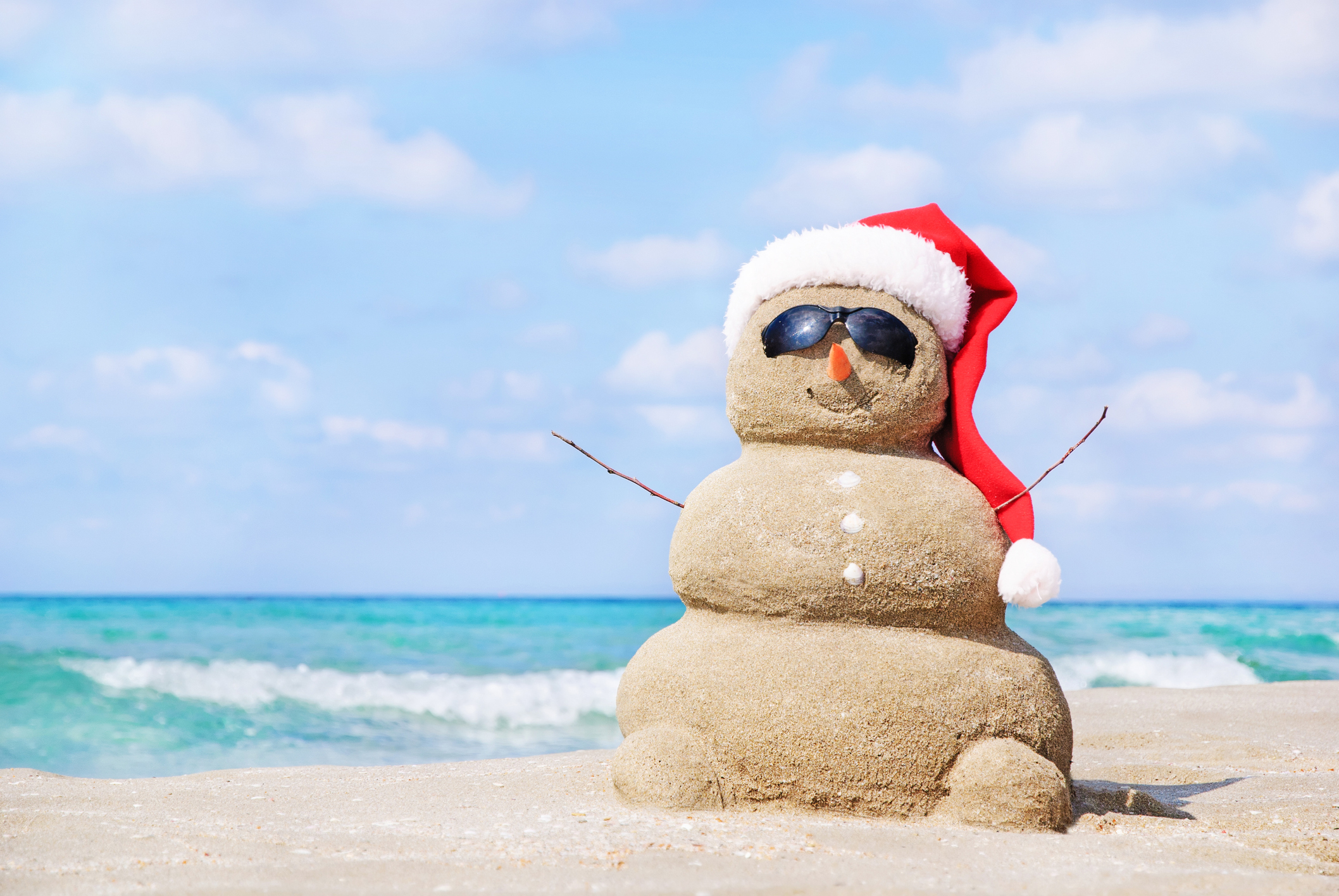 Sandy christmas snowman in red santa hat and sunglasses at sunny beach. Holiday concept for New Years Cards.