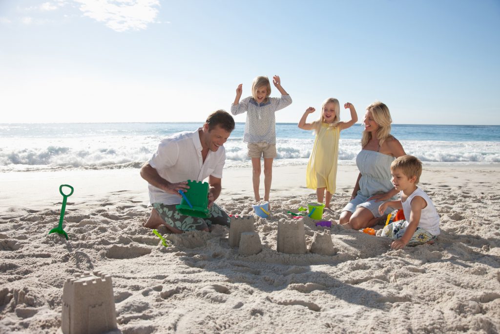 family sitting in sand building a sandcastle with water in the background and sunny day with little clouds making new memories