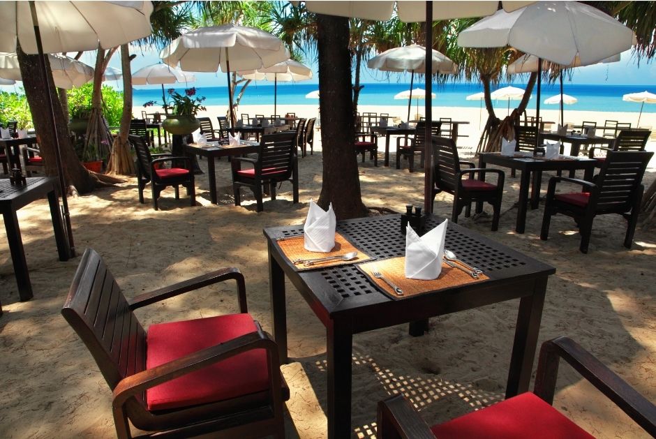 tables and chairs with place settings along with trees and umbrellas on the beach overlooking the blue water