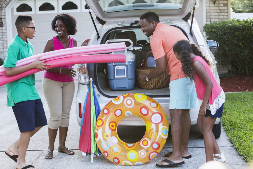 An African American family of four packing their car for a trip to the beach or pool, with cooler, umbrella and toys.  The teenage boy is carrying pool noodles and the 10 year old girl is watching the father put a picnic basket into the car.