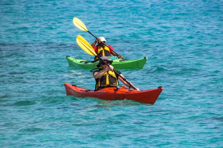 Two kayakers on a green and red kayak each, having an oar with yellow paddles on them, wearing a yellow life vest on calm waters on the gulf of mexico