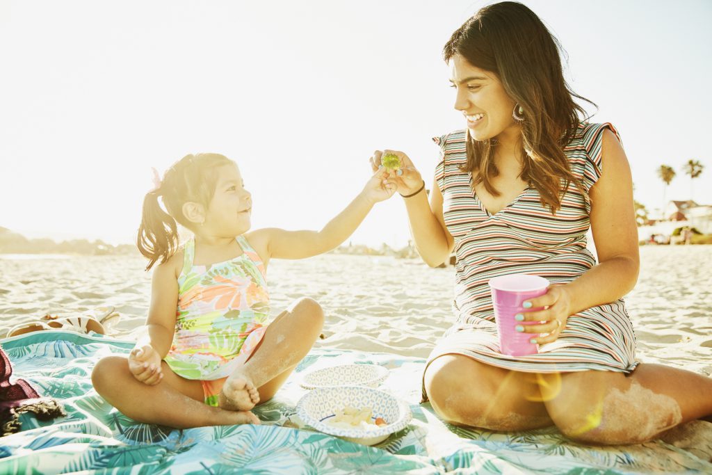 Young girl sharing broccoli with mother while sitting on blanket on beach