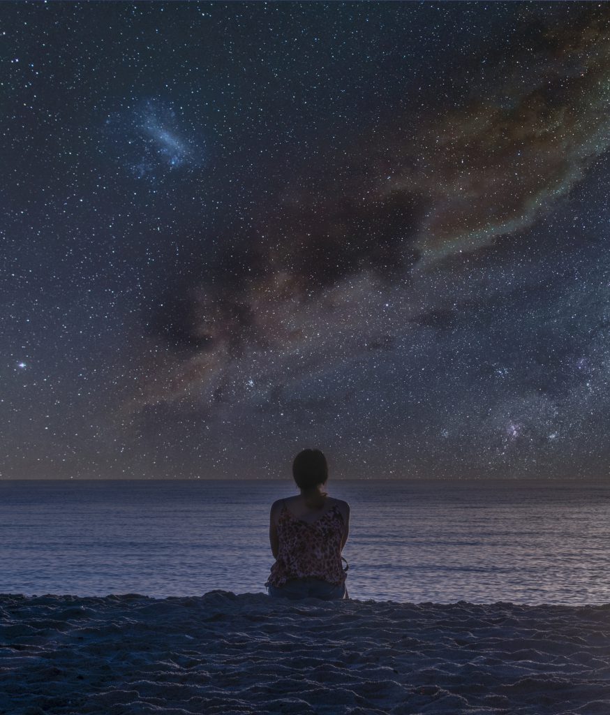 Rear view of a young caucasian woman sitting on a tropical beach watching Milky way in a starry sky